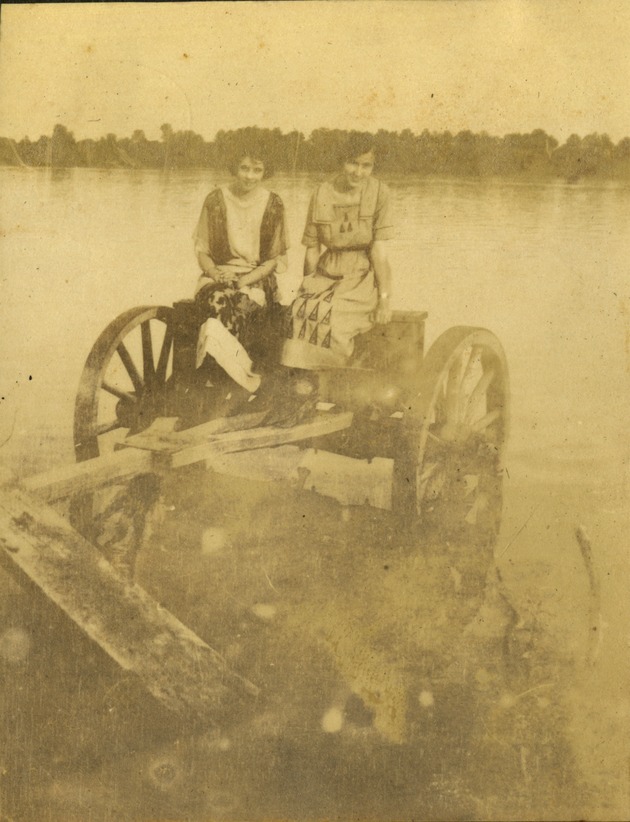 Two women sitting in old carriage wheels by water