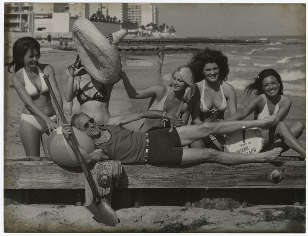 Photographer Chris Hansen relaxing on the beach surrounded by five bathing suit clad models.