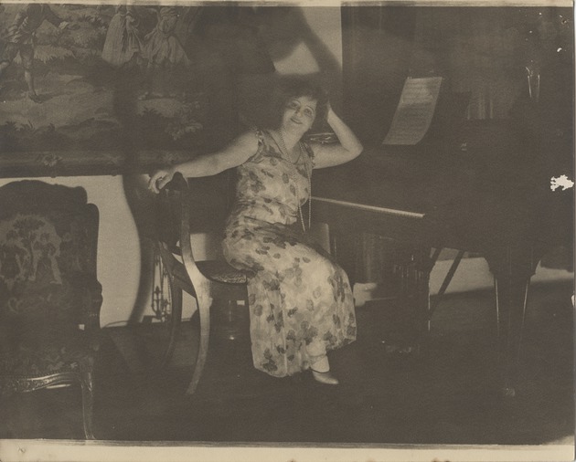 Mana-Zucca pictured in a flower print dress seated at a piano - 