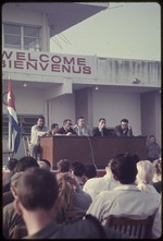 Five men sitting on a stage with a microphone and Cuban flag behind