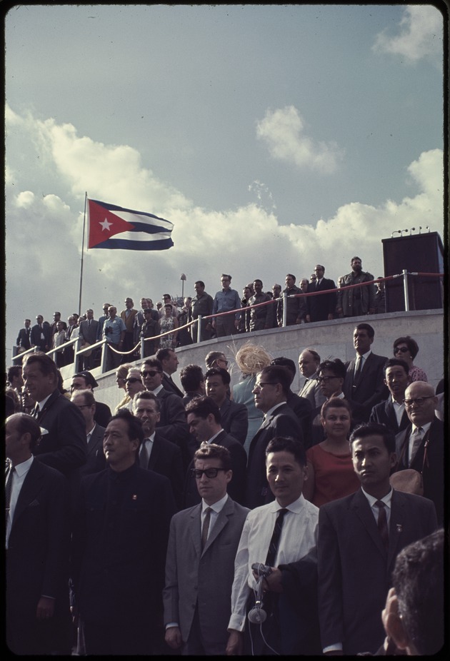 Group of  people standing with Fidel Castro on a stage behind and Cuban flag flying in the distance