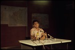 A man sitting at a table with microphones in front of a wall of maps