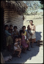 A group of women and children in front of a wooden house with a thatch roof