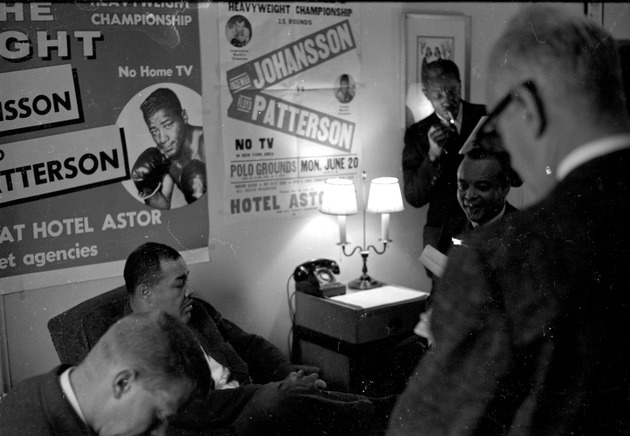 Heavyweight boxer, Joe Louis, sitting in front of Ingemar Johansson and Floyd Patterson fight poster