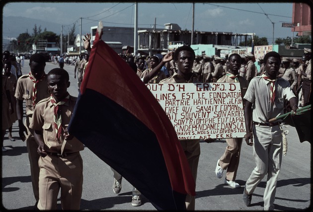 A group of young men carrying flags and a sign marching in the street during the 1990 Haitian general election