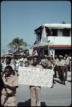 [1990] A young man holding a sign in front of a group of uniformed men on Rue J J Dessalines