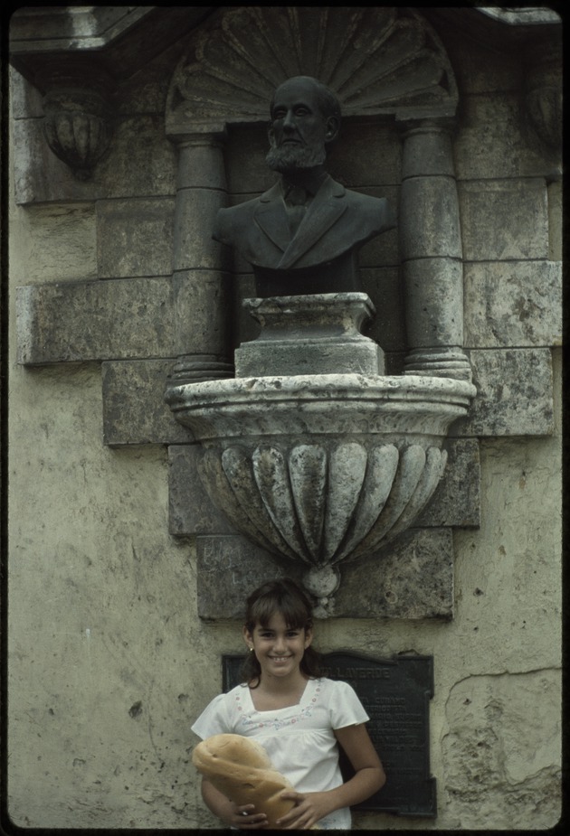 A young girl carrying a loaf of bread standing in front of the Cirilo Villaverde bust