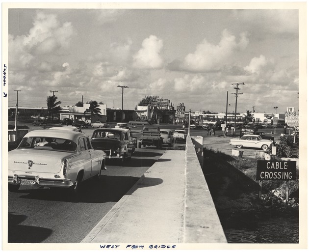 Traffic on John F. Kennedy causeway approaching Normandy Isle - Photograph, recto: [Automobile traffic on bridge west of Normandy Isle. A school on the left, a gas station in the background, and The Sea 'n Sun Motel on the right of bridge]