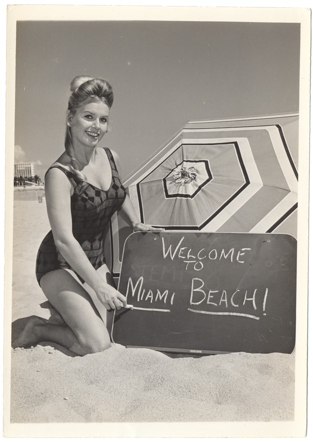 Dolores Kirby next to an umbrellawith a sign that reads "Welcome to Miami Beach" - 