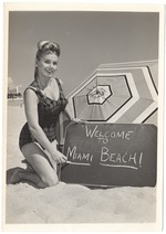 Dolores Kirby next to an umbrellawith a sign that reads Welcome to Miami Beach