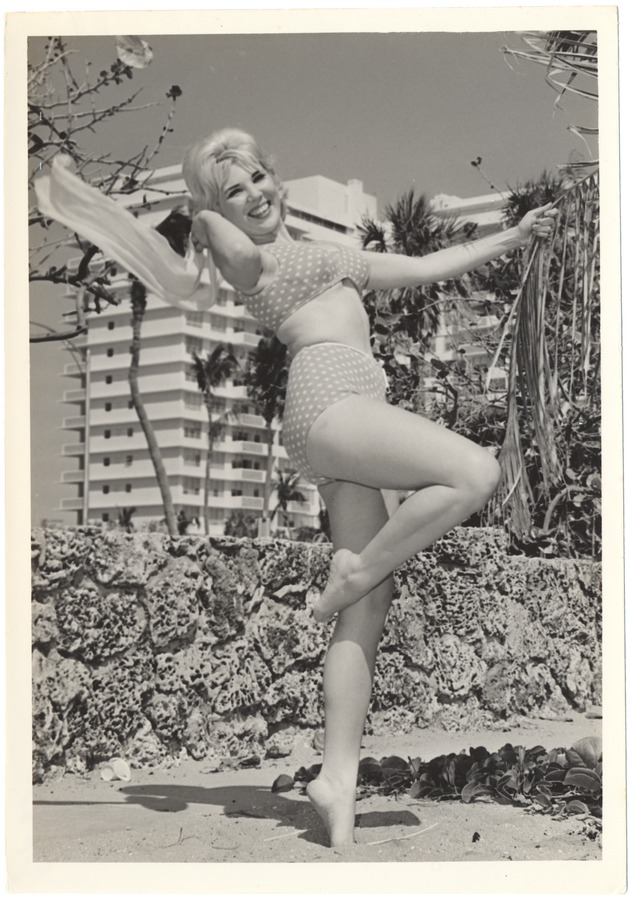 Melinie Smith waving a scarf and grabing a palm tree leaf in front of a coral rock wall - 