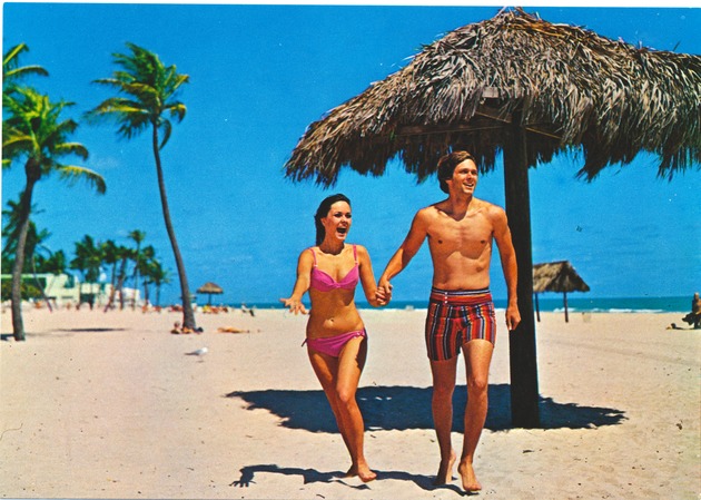 1970s postcards advertising Miami Beach - Postcard, recto: [Couple on the beach with a thatch umbrella and palm trees]