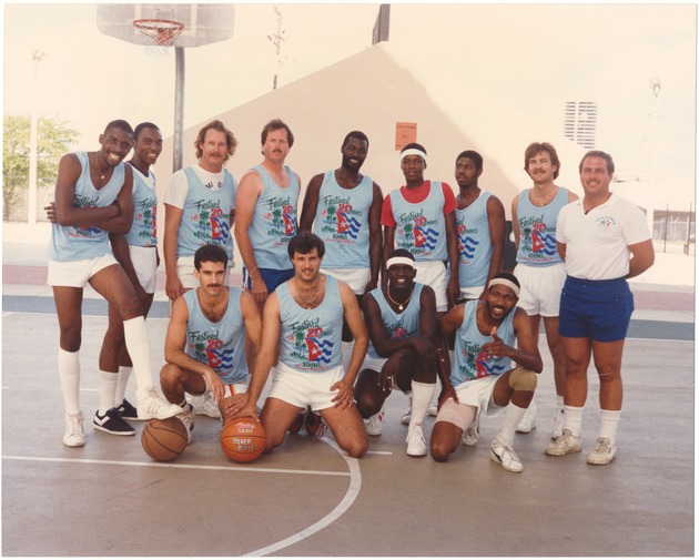 20 de Mayo Festival's basketball tournament - Photograph, recto: [Basketball players celebrating the 20 de Mayo Festival wearing t-shirts featuring Cuban flags and palm trees. Jose Cruz, Miami Beach Budget Office, is second from the right in the back row and Ramon Duenas, Finance Department, is second from the left in the front row, May 20, 1986]