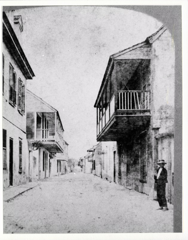 Historic view down St. George Street with Arrivas House on left with balcony, a man standing in front of the De Mesa Sanchez House on right, and a sign reading "BAKERY" hanging from the Avero House on right, looking North