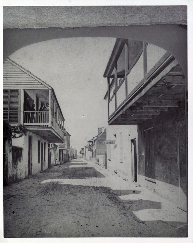 Right side of a stereoview showing a historic view of St. George Street with a group of people on the balcony of the Arrivas House on the left, the De Mesa Sanchez House on the immediate right, and a sign reading "BAKERY" hanging from the Avero House on the right, looking North, ca. 1870-80