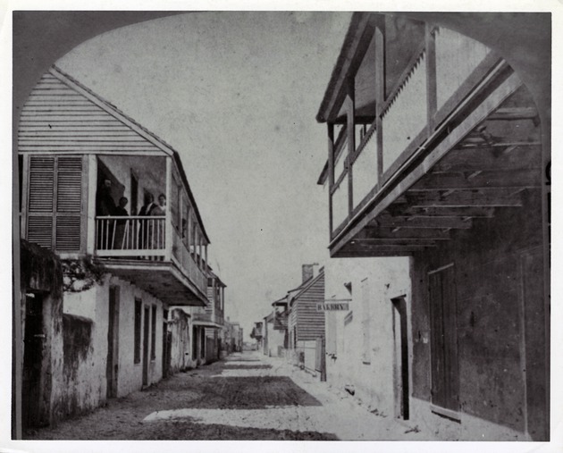 Left side of a stereoview showing a historic view of St. George Street with a group of people on the balcony of the Arrivas House on the left, the De Mesa Sanchez House on the immediate right, and a sign reading "BAKERY" hanging from the Avero House on the right, looking North, ca. 1870-80