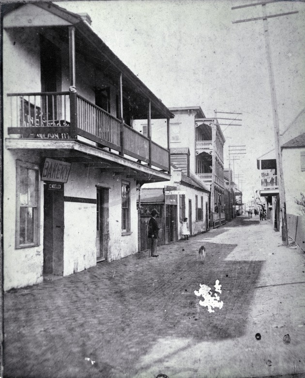 The West side of St. George Street, with a man and a dog in the street in front of the Ortega House, just south of the intersection with Cuna Street with the Columbia Hotel (three strory wood structure with balconies) on the Northwest corner of that intersection, looking north, ca. 1893