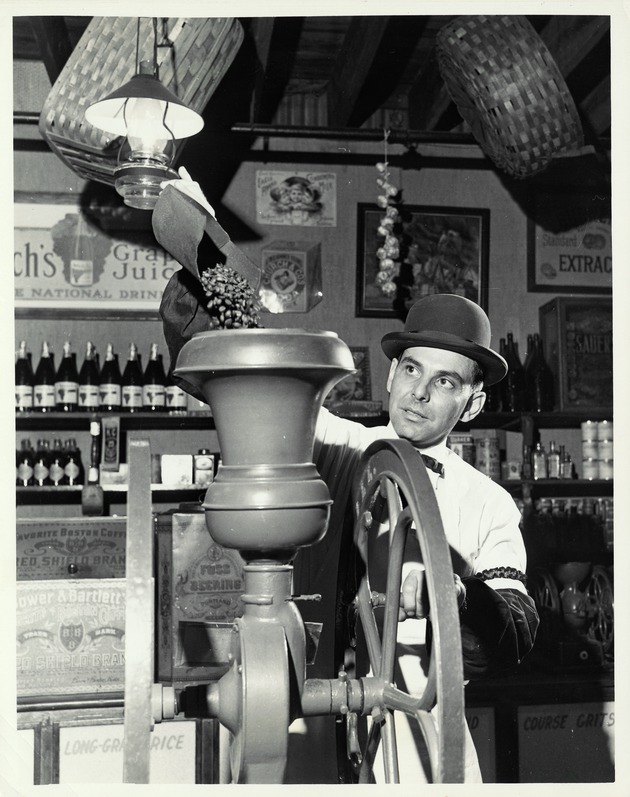 Man in period dress pours whole coffee beans into a hand-cranked grinder in the Old Store Museum, ca. 1965