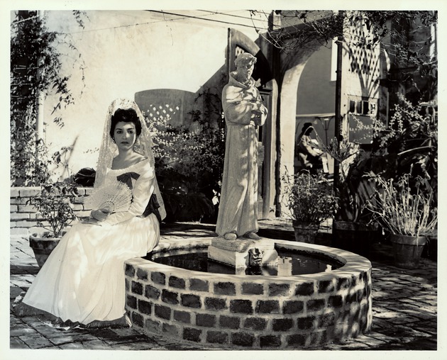 Woman in Spanish period dress sitting by a statue of St. Francis of Assisi in the courtyard of the Oldest House, looking Southeast, ca. 1965