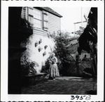 [1960] A woman in period dress in the courtyard on the West side of the Oldest House, looking Southeast, ca. 1960