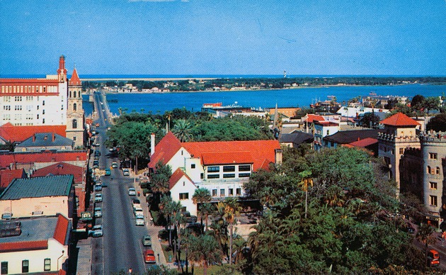 A postcard showing an aerial view of Cathedral Place, the Government House, and the Plaza de la Constitucion, and Anastasia Island beyond, looking East, ca. 1965 - Front