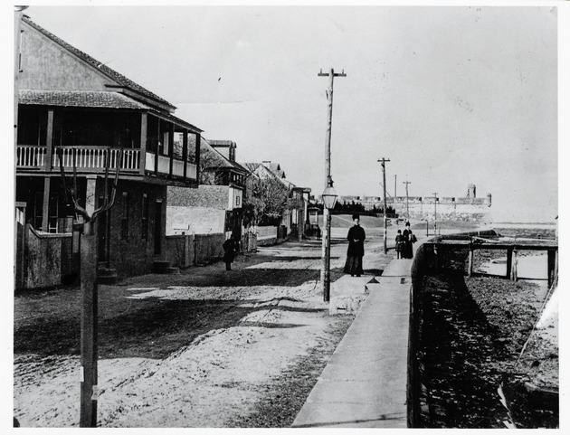 People walking along the seawall along Bay Street (Avenida Menendez) with the Castillo de San Marcos visible in the background, ca. 1880's